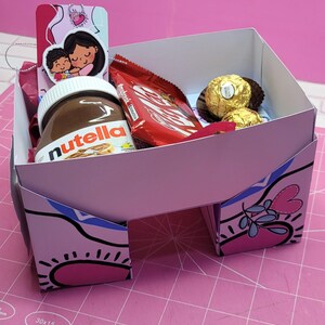 May include: A pink and white cardboard box with a heart design. The box is filled with a jar of Nutella, a chocolate bar, and two chocolate candies. The box is sitting on a pink cutting mat.