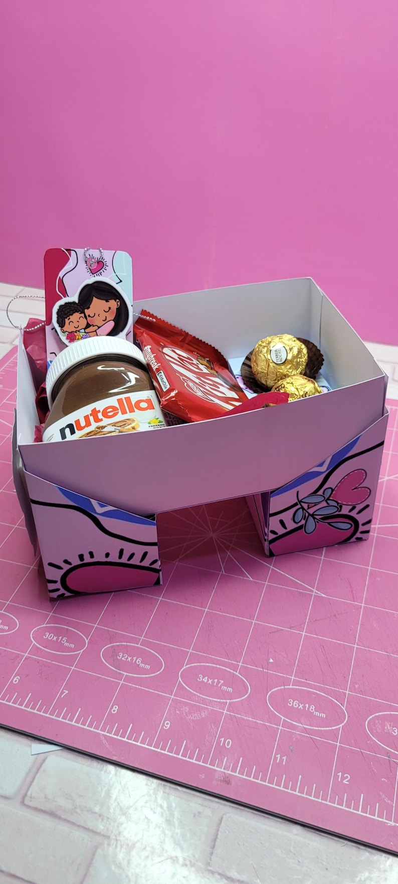 May include: A pink and white cardboard box with a heart design. The box is filled with a jar of Nutella, a chocolate bar, and two chocolate candies. The box is sitting on a pink cutting mat.