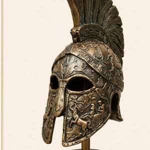 May include: Bronze-colored Spartan helmet with intricate relief carvings of figures and patterns. The helmet features a tall, feathered crest and is mounted on a dark, round base. A decorative piece.