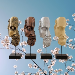 Könnte beinhalten: Vier dekorative Büstenskulpturen eines bärtigen Mannes, jede mit einer anderen metallischen Oberfläche: Gold, Bronze, Silber und Creme. Jede Büste ist auf einem schwarzen quadratischen Sockel mit einem dünnen schwarzen Stab montiert. Der Hintergrund ist ein blauer Himmel mit weißen Blüten.