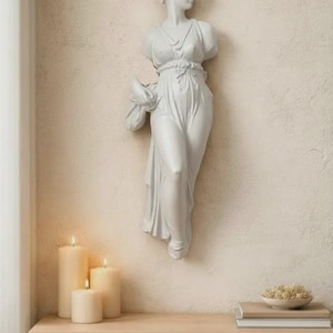 May include: A white plaster wall sculpture of a classical female figure, mounted on a textured beige wall. The figure is draped in flowing robes. Three lit candles and a wooden table with books and a bowl are in the foreground.