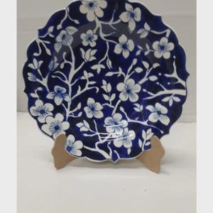 May include: A round ceramic plate with a scalloped edge featuring a deep blue background and a white floral pattern of blossoms and branches. The plate is displayed upright on a small, light-colored wooden stand, showcasing the intricate design.