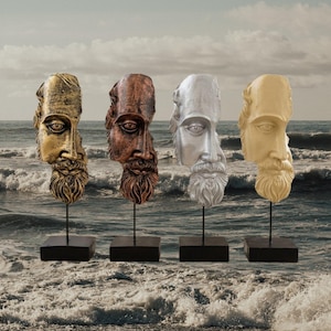 May include: Four bust sculptures of classical figures, each with a different metallic finish: gold, copper, silver, and cream. The sculptures are mounted on black stands, set against a backdrop of a seascape with waves and a cloudy sky.