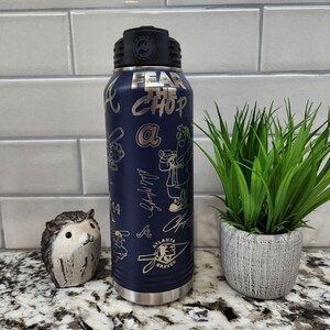 K&ouml;nnte beinhalten: Eine blaue Wasserflasche aus Edelstahl mit schwarzem Deckel und einem wei&szlig;en und goldenen Design. Die Flasche tr&auml;gt die Aufschrift "Fear the Chop" und "Atlanta Braves".