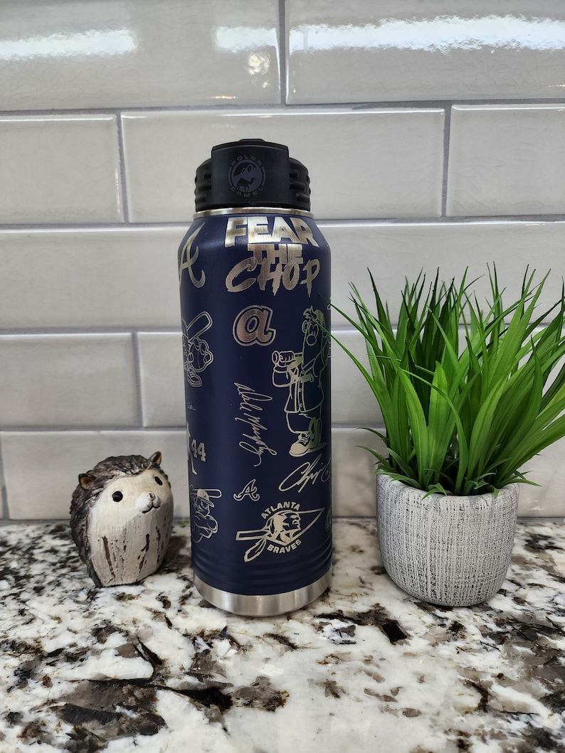 K&ouml;nnte beinhalten: Eine blaue Wasserflasche aus Edelstahl mit schwarzem Deckel und einem wei&szlig;en und goldenen Design. Die Flasche tr&auml;gt die Aufschrift "Fear the Chop" und "Atlanta Braves".
