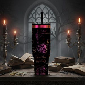 May include: Black tumbler with a pink and black design, featuring a rose, hand gesture, and text that reads "Fuck off I am reading" and "Spread those pages baby". The tumbler has a black straw and is surrounded by candles and books.
