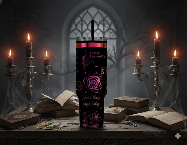 May include: Black tumbler with a pink and black design, featuring a rose, hand gesture, and text that reads "Fuck off I am reading" and "Spread those pages baby". The tumbler has a black straw and is surrounded by candles and books.