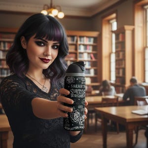 May include: A black and silver water bottle with the text "Sarah's Smutty Book Club" and rose illustrations. The bottle is held by a person in a library setting. The person is wearing a black top and has dark hair with purple highlights.