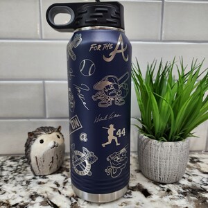 K&ouml;nnte beinhalten: Eine blaue Wasserflasche aus Edelstahl mit schwarzem Deckel und Griff. Die Flasche ist mit einem Baseball-Motiv verziert, darunter ein Baseball, Schl&auml;ger und das Logo der Atlanta Braves. Der Text "For the A" ist in die Flasche graviert.