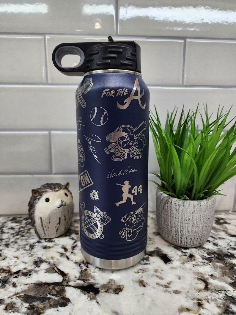 K&ouml;nnte beinhalten: Eine blaue Wasserflasche aus Edelstahl mit schwarzem Deckel und Griff. Die Flasche ist mit einem Baseball-Motiv verziert, darunter ein Baseball, Schl&auml;ger und das Logo der Atlanta Braves. Der Text "For the A" ist in die Flasche graviert.