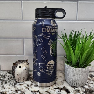 K&ouml;nnte beinhalten: Eine blaue Wasserflasche aus Edelstahl mit schwarzem Deckel und Tragegriff. Die Flasche ist mit einem Baseball-Motiv verziert, darunter ein Cartoon-Baseballspieler, die Worte "World Series Champion 2021" und das Logo der Atlanta Braves.