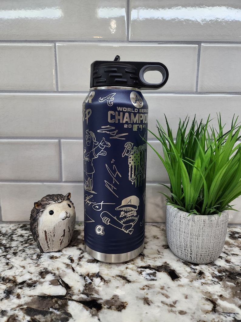 K&ouml;nnte beinhalten: Eine blaue Wasserflasche aus Edelstahl mit schwarzem Deckel und Tragegriff. Die Flasche ist mit einem Baseball-Motiv verziert, darunter ein Cartoon-Baseballspieler, die Worte "World Series Champion 2021" und das Logo der Atlanta Braves.