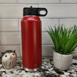K&ouml;nnte beinhalten: Eine rote Edelstahl-Wasserflasche mit schwarzem Deckel und Griff. Die Flasche steht auf einer Granit-Arbeitsplatte.
