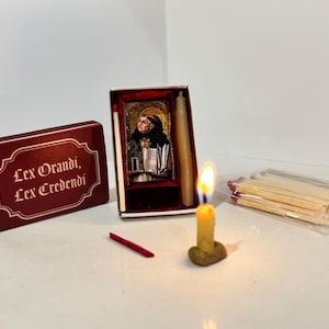 May include: A lit yellow candle with a small flame, next to a box of matches and a small box with an image of a saint. A red box with the text "Lex Orandi, Lex Credendi" is also present.