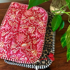 May include: A stack of quilted placemats with floral and animal print designs. The top placemat features a red floral pattern with a striped border. The second placemat has a leopard print design, and the bottom one has black and white stripes.