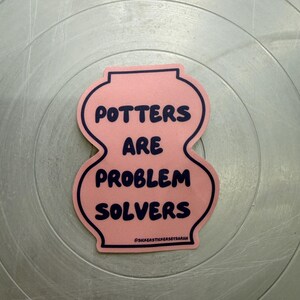 May include: A pink sticker shaped like a vase with a dark blue outline and dark blue text that reads POTTERS ARE PROBLEM SOLVERS. At the bottom, the text reads @SICKERSTICKERSBYSARAH. The sticker has a smooth finish and a distinct curved silhouette.