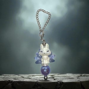 May include: A white fox charm with a silver chain, a blue fan-shaped accent, and a purple bead. The fox is in a seated position with closed eyes. The charm is suspended from a silver chain.