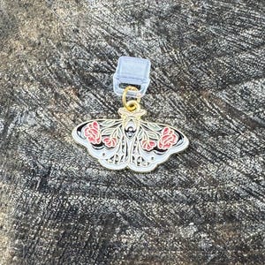 Può includere: Un ciondolo in metallo dorato a forma di falena con dettagli in smalto floreale bianco, nero e rosso sulle ali. Il ciondolo a forma di falena presenta un piccolo anellino dorato fissato a una clip in plastica trasparente per l'aggancio.