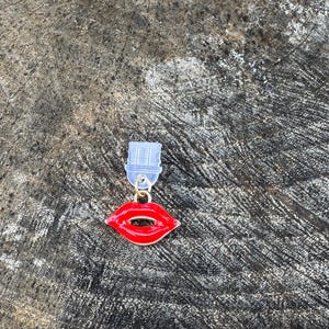 Peut inclure: Une breloque en forme de lèvres en émail rouge fixée à une petite pièce de connexion en plastique transparent. La breloque présente une finition brillante avec un bord en métal doré et une découpe centrale, conçue comme accessoire décoratif.