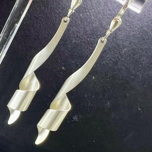Könnte beinhalten: Silberfarbene Hängeohrringe mit abstraktem Design. Jeder Ohrring hat einen langen, gebogenen, flachen Stab, der zu einem spiralförmigen Ende führt. Die Ohrringe werden vor dunklem Hintergrund gezeigt, was ihren metallischen Glanz und ihre besondere Form hervorhebt.