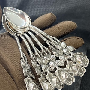Peut inclure: Ensemble de cuillères en argent avec des motifs floraux de roses et de feuilles. Les cuillères sont disposées en éventail, mettant en valeur leur finition polie et les manches en forme de branches, témoignant d'un travail artisanal.
