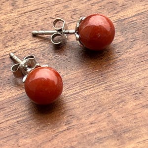 Op de afbeelding: Een paar oorstekers met ronde, rood-oranje edelstenen. De oorbellen hebben zilverkleurige pinnen en sluitingen. De oorbellen worden gepresenteerd op een houten oppervlak.