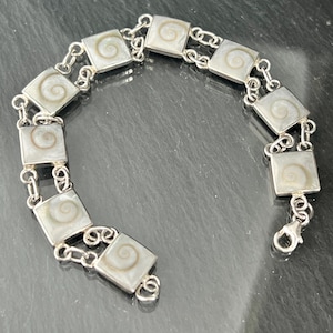 Może przedstawiać: Srebrna bransoletka z kwadratowymi, białymi zawieszkami z muszli z motywem spirali. Bransoletka ma zapięcie na karabińczyk.