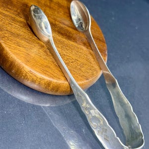 Könnte beinhalten: Ein Paar silberfarbene Zuckerzange aus Metall mit löffelförmigen Enden und einem eingravierten dekorativen Muster am Griff. Die Zange hat ein geschwungenes, flexibles Design, das die beiden Arme an einer abgerundeten Basis verbindet.