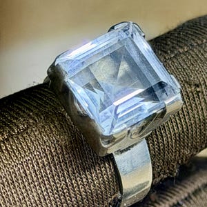 Op de afbeelding: Een zilveren ring met een grote, heldere, vierkant geslepen edelsteen. De edelsteen is gezet in een zilveren band met decoratieve details. De ring wordt gepresenteerd op een bruin stoffen oppervlak.