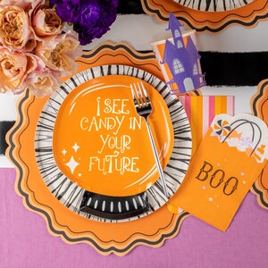 Puede incluir: Decoraciones para fiestas de Halloween. Un plato naranja con el texto "I SEE CANDY IN YOUR FUTURE" y un tenedor plateado. Incluye una bolsa de golosinas "BOO", una taza de casa embrujada morada y platos decorativos con rayas blancas y negras.