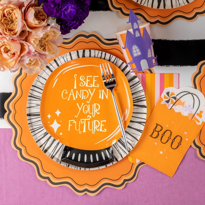 Puede incluir: Decoraciones para fiestas de Halloween. Un plato naranja con el texto "I SEE CANDY IN YOUR FUTURE" y un tenedor plateado. Incluye una bolsa de golosinas "BOO", una taza de casa embrujada morada y platos decorativos con rayas blancas y negras.
