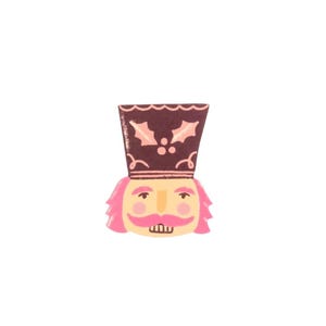 May include: A whimsical illustration of a nutcracker soldier. The nutcracker has a light yellow face, pink hair and mustache, and a dark brown hat decorated with pink holly leaves and berries. The design is set against a white background.