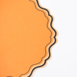 Puede incluir: Un elemento de papel naranja con un borde festoneado y un borde negro y naranja claro. El dise&ntilde;o se establece sobre un fondo blanco, creando un contraste. La forma recuerda a una etiqueta decorativa.