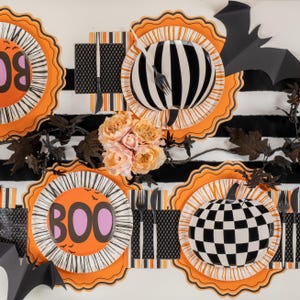 Puede incluir: Decoraci&oacute;n de mesa de Halloween con platos naranjas, negros y blancos. Los platos presentan dise&ntilde;os como rayas, cuadros y la palabra "BOO". Cubiertos negros y murci&eacute;lagos de papel se suman a la decoraci&oacute;n festiva. Un ramo de flores color melocot&oacute;n y hojas oscuras completan el arreglo.