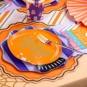 Puede incluir: Una mesa decorada con tem&aacute;tica de Halloween con detalles en naranja, morado y blanco. Un plato naranja con el texto "I SEE CANDY IN YOUR FUTURE" se combina con un tenedor plateado. Otros elementos incluyen una taza con forma de casa encantada y abanicos de papel decorativos.