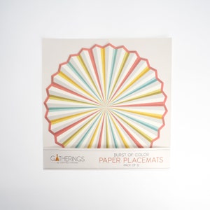May include: A pack of 12 paper placemats with a burst of color design. The placemats are round and have a radial pattern of pink, yellow, and blue stripes. The text "BURST OF COLOR PAPER PLACEMATS PACK OF 12" is printed on the packaging.