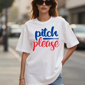 Pode incluir: Uma t-shirt branca de manga curta com o texto "pitch please" impresso na frente. A palavra "pitch" está em azul com um gráfico de um taco de basebol por baixo, e a palavra "please" está em vermelho, ambas em letra cursiva.