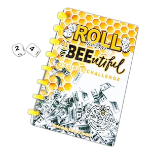 May include: A savings challenge notebook with a yellow honeycomb design and the words "Roll the Dice BEEautiful Savings Challenge." The notebook features a cartoon bee and illustrations of dice and money. Two dice with numbers 2 and 4 are next to the notebook.