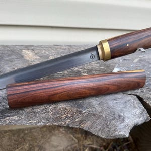 May include: A fixed-blade knife with a dark, textured metal blade featuring a small shield-shaped logo. The handle is crafted from dark brown wood with a brass bolster. A matching wooden sheath lies horizontally in front of the knife blade.