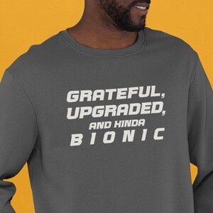 May include: A dark gray sweatshirt with white text that reads "GRATEFUL, UPGRADED, AND KINDA BIONIC".