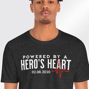 May include: A dark grey t-shirt with the text "POWERED BY A HERO'S HEART" in white and a red heartbeat graphic. The date "02.08.2020" is printed below the text.