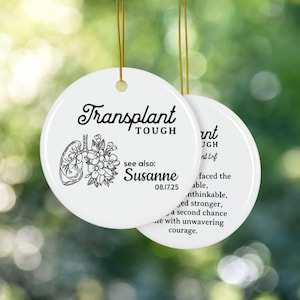 Può includere: Ornamento rotondo in ceramica bianca con cordino dorato. Presenta il testo "Transplant Tough", un'illustrazione di polmoni e fiori, il nome "Susanne" e la data "08.17.25".