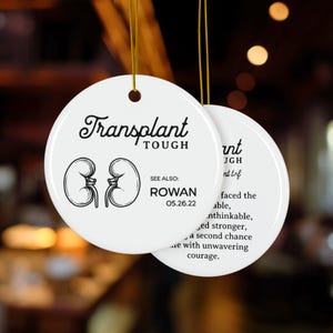 Puede incluir: Adorno redondo de cerámica blanca con el texto "Transplant Tough" y una ilustración de riñón. El adorno también incluye el nombre "Rowan" y la fecha "05.26.22". Cuelga de un hilo dorado.