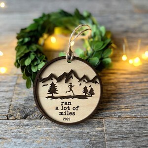 May include: A circular wooden ornament featuring a runner, trees, and mountains. The text "ran a lot of miles 2025" is engraved on the light wood. It has a dark brown border and a twine loop for hanging. The ornament is approximately 8 cm in diameter.