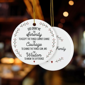 Op de afbeelding: Twee witte keramische ornamenten met gouden ophangkoorden. Eén ornament heeft de tekst "God Grant Me the Serenity..." in zwart schrift, omgeven door een groene en rode bloemenrand. Het tweede ornament heeft het woord "Family".