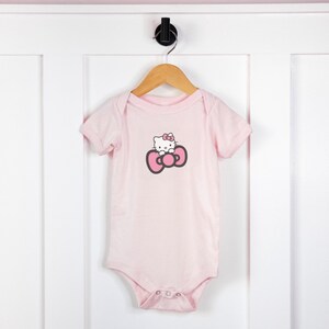 Op de afbeelding: Een roze baby-romper met een Hello Kitty-afbeelding op de voorkant. De afbeelding toont Hello Kitty met een roze strik.
