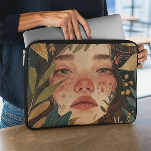 May include: A laptop sleeve featuring a colourful illustration of a woman's face surrounded by green and brown leaves. The woman has green eyes and freckles.