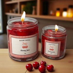 May include: Two glass jar candles with lit wicks, filled with red wax, and labeled "Sugar Candle Cherry Jam." The labels have a gingham border and cherry illustration. Fresh cherries are scattered on the wooden surface.