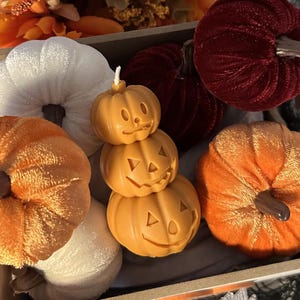 May include: A set of three stacked pumpkin-shaped candles with jack-o'-lantern faces. The candles are surrounded by various fall-themed decorations, including velvet pumpkins and a white pumpkin.