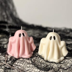 May include: Two ghost-shaped candles, one pink and one white, with black eyes, sitting on a black lace surface.